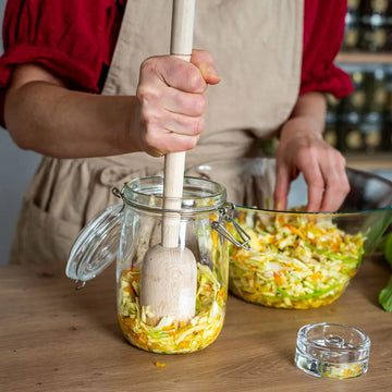 Eine Person drueckt mit einem Holzstampfer geschnittenes Gemuese in ein 1-Liter-Drahtbuegelglas, daneben steht eine Glas Schuessel mit frischem Kraut.