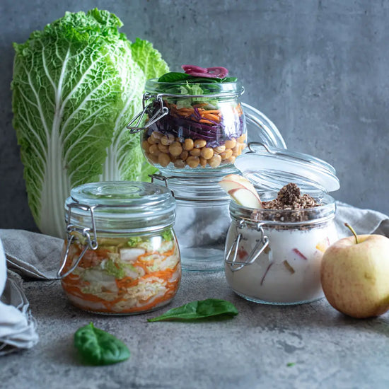 Drei 500-ml-Buegelglaeser sind gefuellt mit Salat, Kimchi und veganem Fruehstueck; dahinter stehen Chinakohl und ein frischer Apfel.
