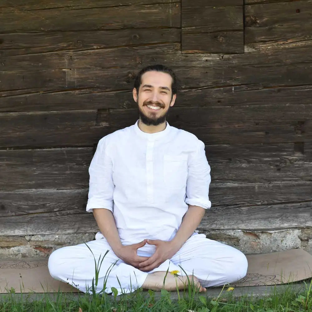 Mann in weißer Kleidung sitzt meditierend im Schneidersitz vor einer rustikalen Holzwand. Symbolbild für Achtsamkeit, Meditation, Stressabbau, innere Balance und ganzheitliche Gesundheit in der Natur.