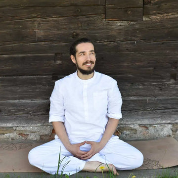 Naturverbundener Mann in weißer Kleidung sitzt entspannt im Schneidersitz auf einer Yogamatte vor einer rustikalen Holzwand. Die ruhige Szene vermittelt Achtsamkeit, Meditation, innere Balance und einen ganzheitlichen Lebensstil in der Natur.