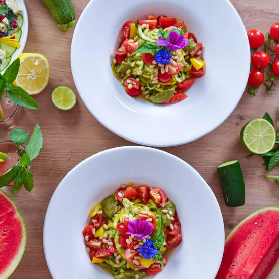 Zwei Schalen Zucchininudelsalat mit Tomaten, Pinienkernen und Blüten, umgeben von Limetten und Wassermelone.
