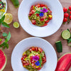 Zwei Schalen Zucchininudelsalat mit Tomaten, Pinienkernen und Blüten, umgeben von Limetten und Wassermelone.