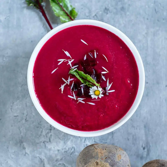 Cremige vegane Rote-Bete-Suppe in einer weißen Schüssel, garniert mit frischer Roter Bete, essbaren Blüten und Kräutern. Nährstoffreiche, vollwertige Gemüsesuppe für eine gesunde pflanzliche Ernährung.