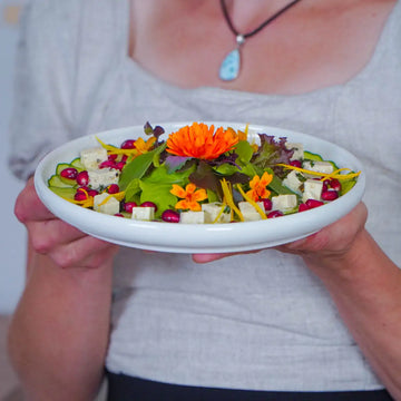 Frau hält Teller mit Blattsalat, Fetawürfeln, Granatapfelkernen, Gurke und essbaren Blüten dekoriert.