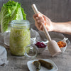 Ein 1-Liter-Fermentationsglas mit Sauerkraut steht neben einem Glasgewicht, waehrend eine Hand einen Holzstampfer haelt; im Hintergrund liegen weitere Fermente.