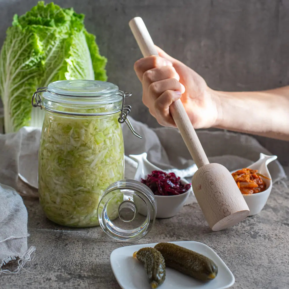 Ein 1-Liter-Fermentationsglas mit Sauerkraut steht neben einem Glasgewicht, waehrend eine Hand einen Holzstampfer haelt; im Hintergrund liegen weitere Fermente.