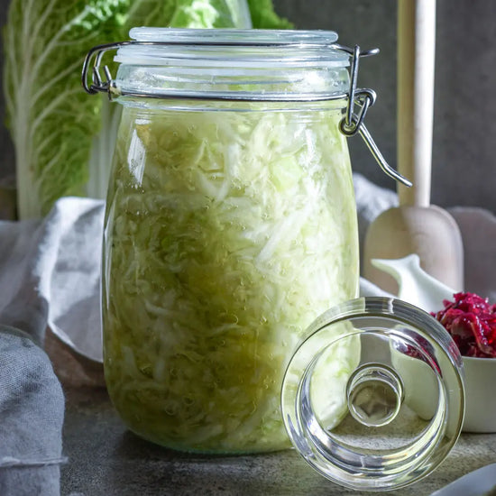 Ein 1-Liter-Drahtbuegelglas ist mit frischem Sauerkraut gefuellt, daneben liegt ein Glasgewicht und im Hintergrund stehen Kohl und ein Holzstampfer.