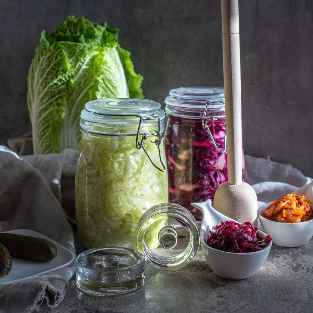 Ein 1-Liter-Fermentationsglas mit Sauerkraut steht neben Rotkohlferment, Glasgewichten, einem Holzstampfer und frischem Chinakohl.