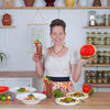 Frau lächelt und präsentiert halbe Wassermelone und Drink, davor Tisch mit Salattellern und Gemüse.
