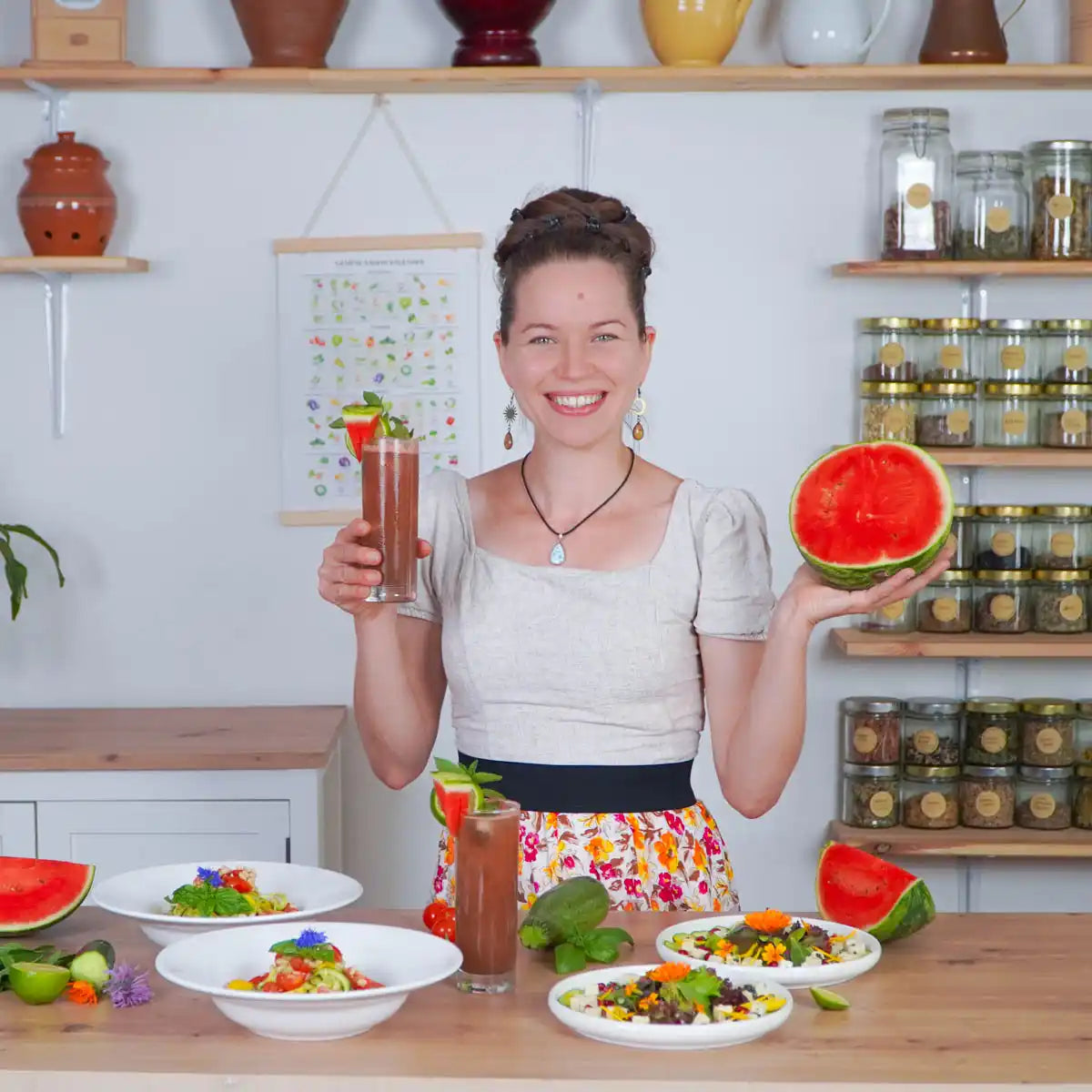 Frau lächelt und präsentiert halbe Wassermelone und Drink, davor Tisch mit Salattellern und Gemüse.