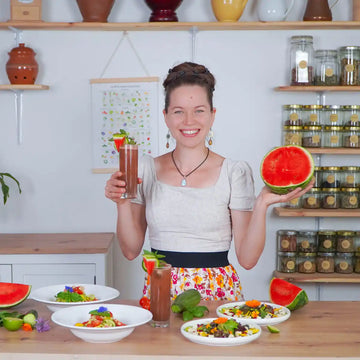 Frau lächelt und präsentiert halbe Wassermelone und Drink, davor Tisch mit Salattellern und Gemüse.