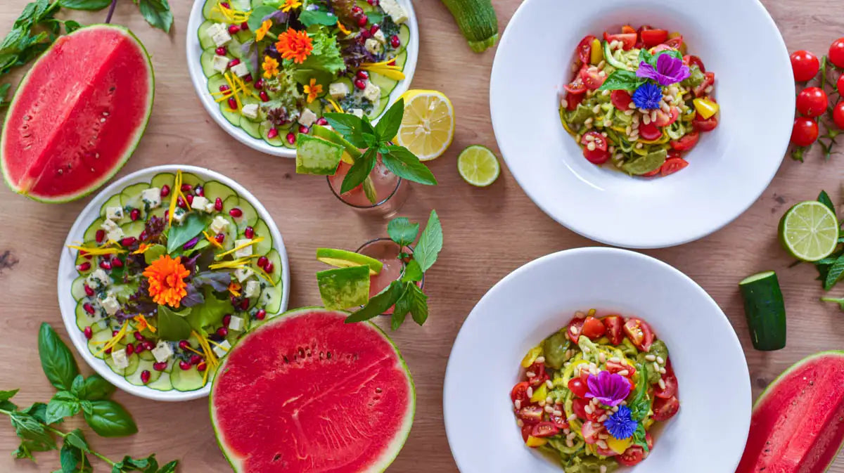 Vier Salatteller mit Zucchininudeln und Gurken-Carpaccio, dazwischen Wassermelone, Drinks und Kräuter.