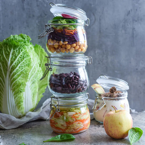 Drei gefuellte 500-ml-Drahtbuegelglaeser sind gestapelt, gefuellt mit Gemuese, Bohnen und Fermenten; daneben stehen Obst und ein weiteres Glas.