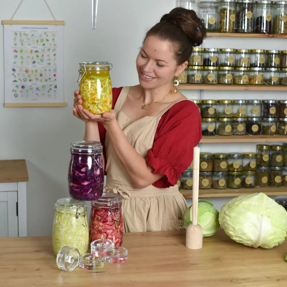 Theresa hält ein Glas bunt fermentiertes Gemüse, vor ihr stehen weitere Fermentationsgläser, Glasgewichte und frischer Kohl.