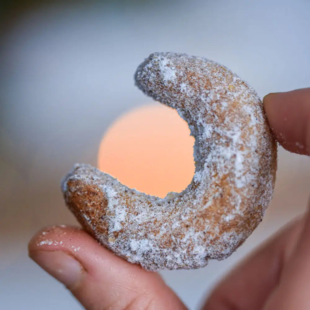 Ein Vanillekipferl mit Puderzucker wird zwischen zwei Fingern gehalten, im Hintergrund ein warmes unscharfes Licht.