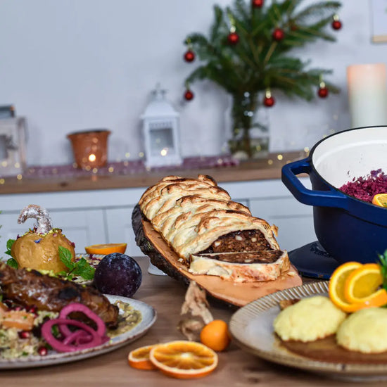 Vegane Festtagstafel mit Nussbraten im Blätterteig, Rotkraut, Knödeln und weihnachtlicher Dekoration im Hintergrund.