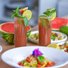 Zwei Gläser Wassermelonen-Drink mit Minze und Limette im Vordergrund, dahinter Salate und Wassermelone.