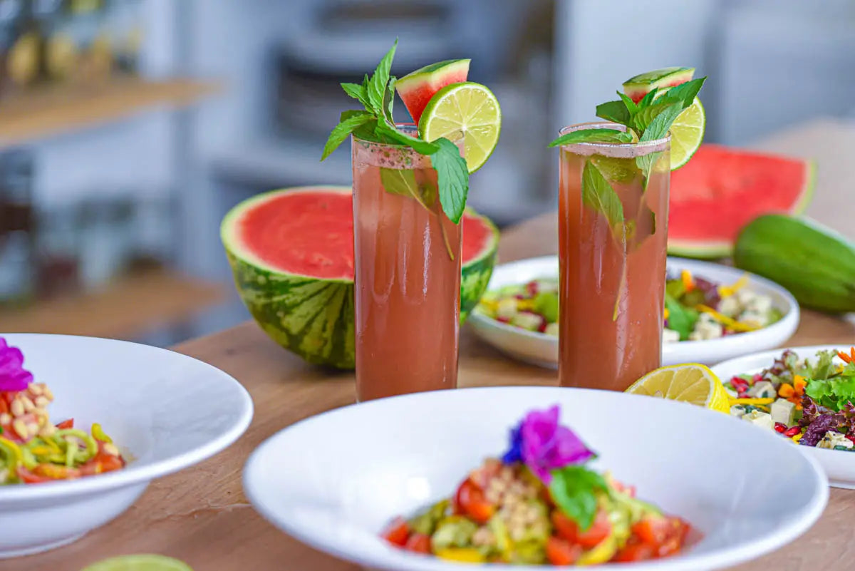 Zwei Wassermelonen-Drinks mit Limette und Minze auf gedecktem Tisch, umgeben von Zucchininudel- und Carpaccio-Salaten.