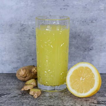 Glas mit hellgelbem Zitronen-Ingwer-Detox-Getränk auf grauer Steinoberfläche. Daneben liegen frische Ingwerstücke und eine halbierte Zitrone – ideal für natürliche Entgiftung und Erfrischung.