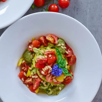 Draufsicht auf Schale mit Zucchininudeln, Cherrytomaten, Pinienkernen und Kornblume, umgeben von frischen Tomaten.