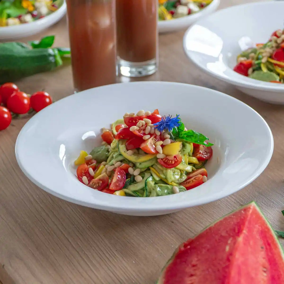 Schale mit Zucchininudeln, Cherrytomaten und Pinienkernen, im Hintergrund Drinks, weitere Salate und Wassermelone.