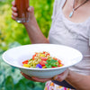 Frau hält Schale mit Zucchininudeln, Tomaten und Pinienkernen sowie ein Glas Getränk im Garten.