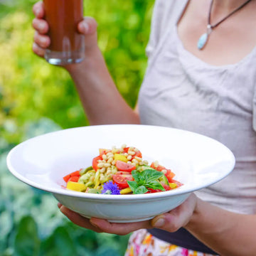Frau hält Schale mit Zucchininudeln, Tomaten und Pinienkernen sowie ein Glas Getränk im Garten.